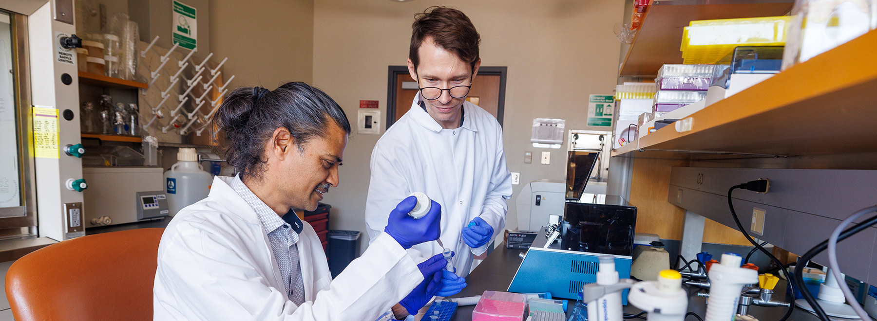 A standing researcher holds a test tube while a sitting researcher uses a pipette to fill the tube